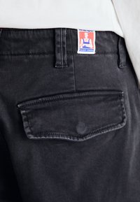 Schwarze Jeans aus Denim verfügen über eine Hintertasche mit einer abgerundeten Klappe und sichtbaren Nähten. Das Etikett zeigt "Wrangler" mit einer Cartoonfigur.