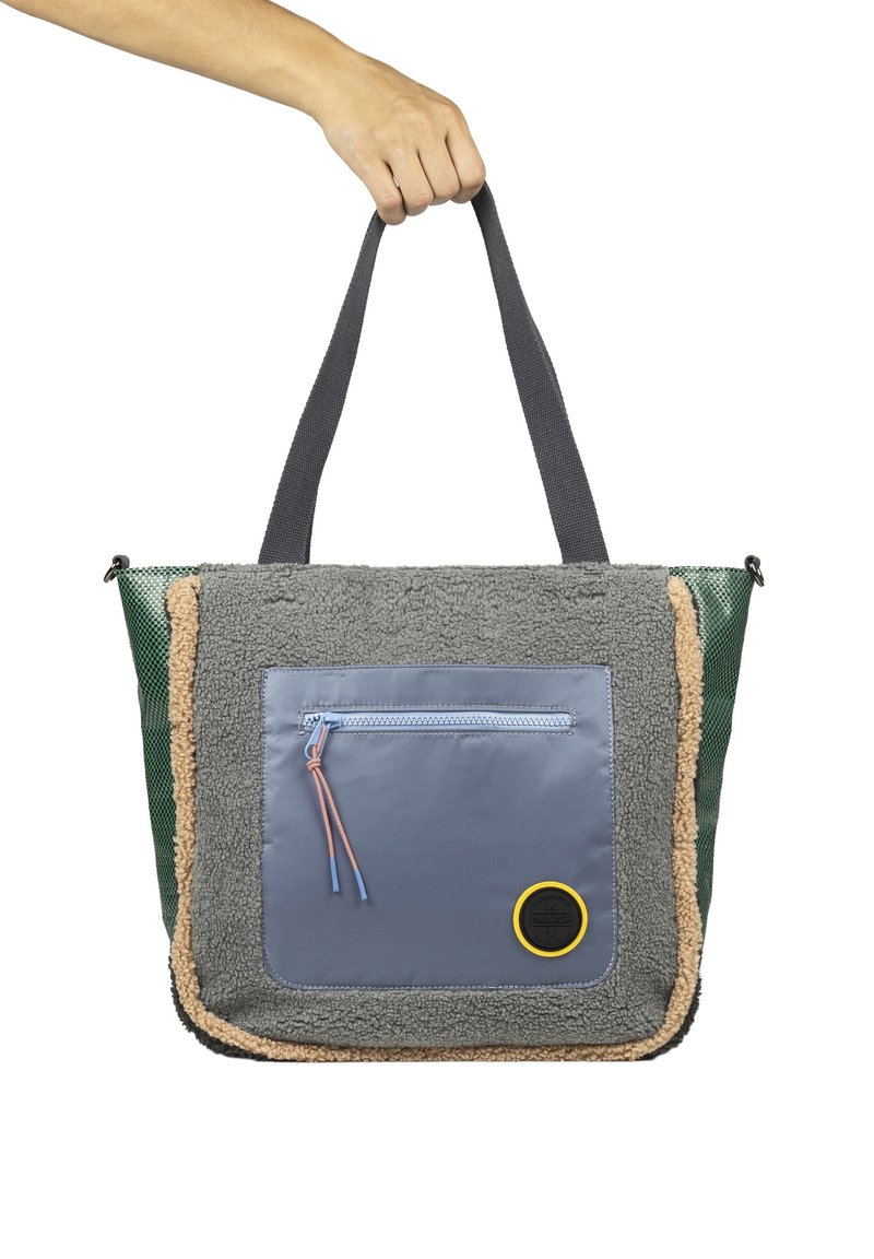 Bolsa de mano multicolor con felpa gris texturizada, bolsillo delantero de nailon azul, lados verdes y acento circular amarillo. Asas de tela gris resistente.
