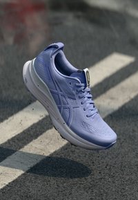 Gaiši purpura skriešanas apavi ar teksturētu virsu, acs dizainu un baltu zoli. Iekļauj šņorēšanas elementus un zīmola akcentus, kas nodrošina stabilitāti.