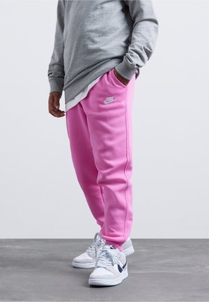 Person trägt leuchtend pinke Nike-Sweathosen, graues Sweatshirt und weiße Turnschuhe mit marineblauen und hellblauen Details, steht auf grauem Boden.