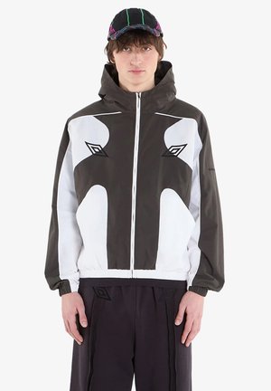 Giovane uomo che indossa una giacca nera e bianca con zip e motivi geometrici e un cappellino nero con cuciture al neon, in piedi davanti a uno sfondo uniforme.