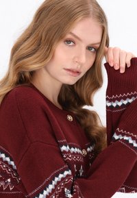 Bordó kötött pulóver arany gombokkal, fehér és tengerész kék mintás díszítéssel az ujjakon, bordázott mandzsettákkal és lazább szabással.