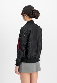 Svart bomberjakke med ribbestrikkede mansjetter og kant, sidelommer og en fremtredende rød merkedetalj på armen. Kombinert med et grått skjørt.