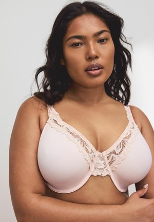 Woman with medium-dark skin and wavy dark hair wearing a light pink lace-trimmed bra, looking directly ahead with a neutral expression.