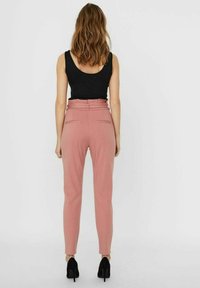 Pantalon moulant rose taille haute, à texture lisse, avec deux poches arrière, associé à un haut noir sans manches et des talons noirs.