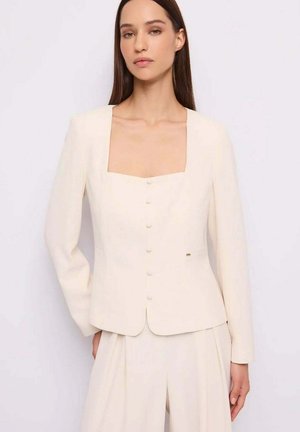 Donna che indossa una giacca su misura color avorio con bottoni coperti e pantaloni a gamba larga abbinati, su uno sfondo chiaro e uniforme.