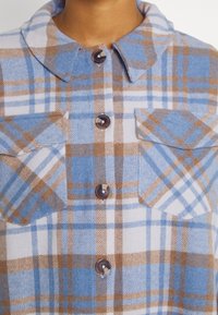 Primo piano di una camicia a quadri blu, bianca e marrone con colli piegati, due tasche sul petto e bottoni rotondi scuri.