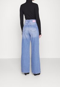 Donna che indossa jeans a vita alta e a gamba larga di colore azzurro chiaro e un top nero fitted a maniche lunghe, in piedi di spalle su uno sfondo bianco uniforme.