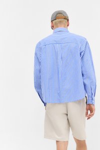 Mand iført en blåt og hvidt stribet langærmet skjorte, beige shorts og en gråternet kasket, som vender væk, på en hvid baggrund.