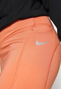 Nike legíny v živé oranžové barvě, vyrobené z hladkého materiálu, s plochými švy a jemným stříbrným logem na stehně.