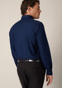 Granatowa koszula z długim rękawem i usztywnianym kołnierzem. Koszula ma gładką fakturę i jest zestawiona z ciemnymi spodniami oraz czarnym paskiem.