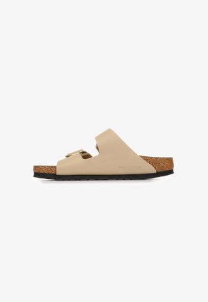 Beige Birkenstock slidesandaal met twee verstelbare banden, kurken voetbed en zwarte gestructureerde zool, gezien van de zijkant op een witte achtergrond.