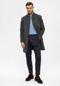 Grauer Wollmantel mit Stehkragen, getragen über einem hellblauen Hemd mit Knopfleiste. Dunkle Hose und braune Wildlederschuhe vervollständigen das Outfit.