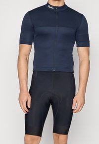 Camiseta de ciclismo azul marino con mangas cortas, cremallera frontal y logo de Rapha; combinada con pantalones cortos de ciclismo acolchados negros con puños elásticos.