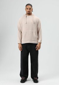 Beige fleece hoodie met een voorzak en trekkoorden, gecombineerd met losse zwarte jeans en zwarte schoenen met roze veters, tegen een grijze achtergrond.