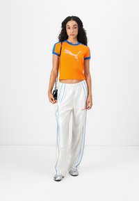 Pomarańczowa skrócona bluzka z niebieskimi akcentami i logo PUMA. Białe luźne spodnie z niebieskimi paskami i pomarańczowym napisem. Srebrne buty.