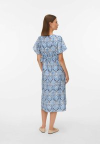 Mujer con cabello lacio castaño de pie de perfil, vistiendo un vestido midi azul con estampado, de mangas cortas y zapatos planos beige.