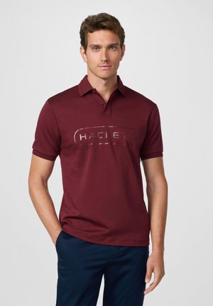 Camisa polo burdeos con cuello y mangas cortas, que presenta un gran logo en relieve en la parte frontal. Textura de tela suave, diseño simple.