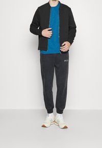 Giacca bomber nera con zip, T-shirt blu strutturata, pantaloni della tuta scuri con logo e sneakers chiare con dettagli blu.