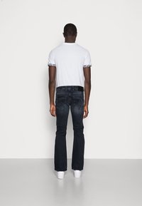 Mężczyzna stojący tyłem, ubrany w białą koszulkę z kwiatowym obszyciem na rękawach, ciemne dżinsy i białe trampki na jednolitym tle.