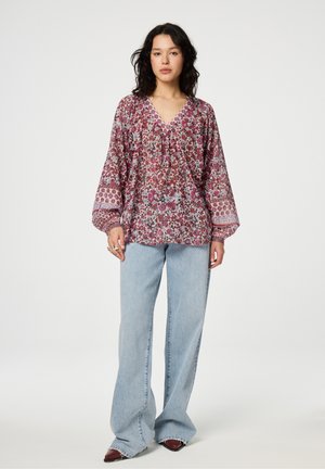 Bluse mit Blumenmuster, V-Ausschnitt und langen Ärmeln, mit Raffungen. Kombiniert mit lockeren, hellblauen Jeans und roten Schuhen.