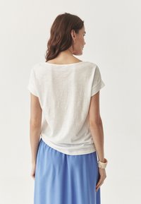 Hvid kortærmet top med scoop halsudskæring, lavet af letvægtsstof, med en afslappet pasform. Sammen med en blå nederdel og et armcuff.