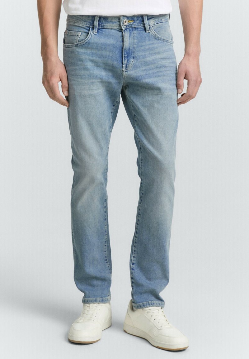 Man in lichtblauwe slim-fit jeans en witte sneakers, staand tegen een effen achtergrond.