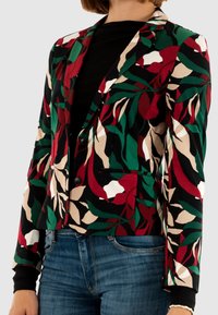 Vrouw die een op maat gemaakte blazer draagt met abstracte bladmotieven in rood, groen, beige en zwart over een zwarte top en blauwe jeans.