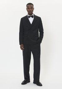 Czarna marynarka smokingowa z podwójnym zapięciem i satynowymi klapami, czarne spodnie, biała koszula i czarny muszka. Lśniące czarne buty oksfordki. Jednolity tło.