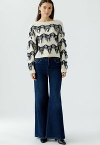 Strickpullover in Creme mit navyfarbenen Fransen, kombiniert mit hochgeschnittenen, ausgestellten blauen Jeans. Flache schwarze Stiefeletten runden den Look ab.
