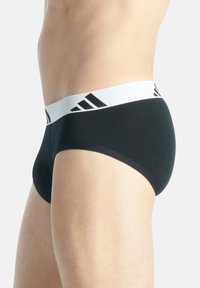 Schwarze Baumwollslips mit einem weißen elastischen Bund, der mit schwarzen Adidas-Logo-Streifen versehen ist. Glatte Textur und körpernahe Passform.