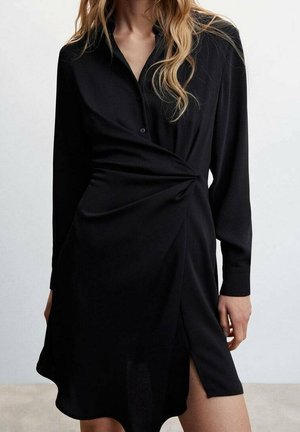 Frau trägt ein langärmeliges schwarzes Kleid mit gerafftem Detail an der Taille und einem Schlitz vorne, steht vor einem schlichten Hintergrund.