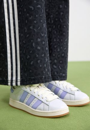 Baskets blanches avec des rayures lavande portées avec un pantalon noir avec des bandes blanches sur les côtés et un motif léopard subtil, debout sur un sol vert.