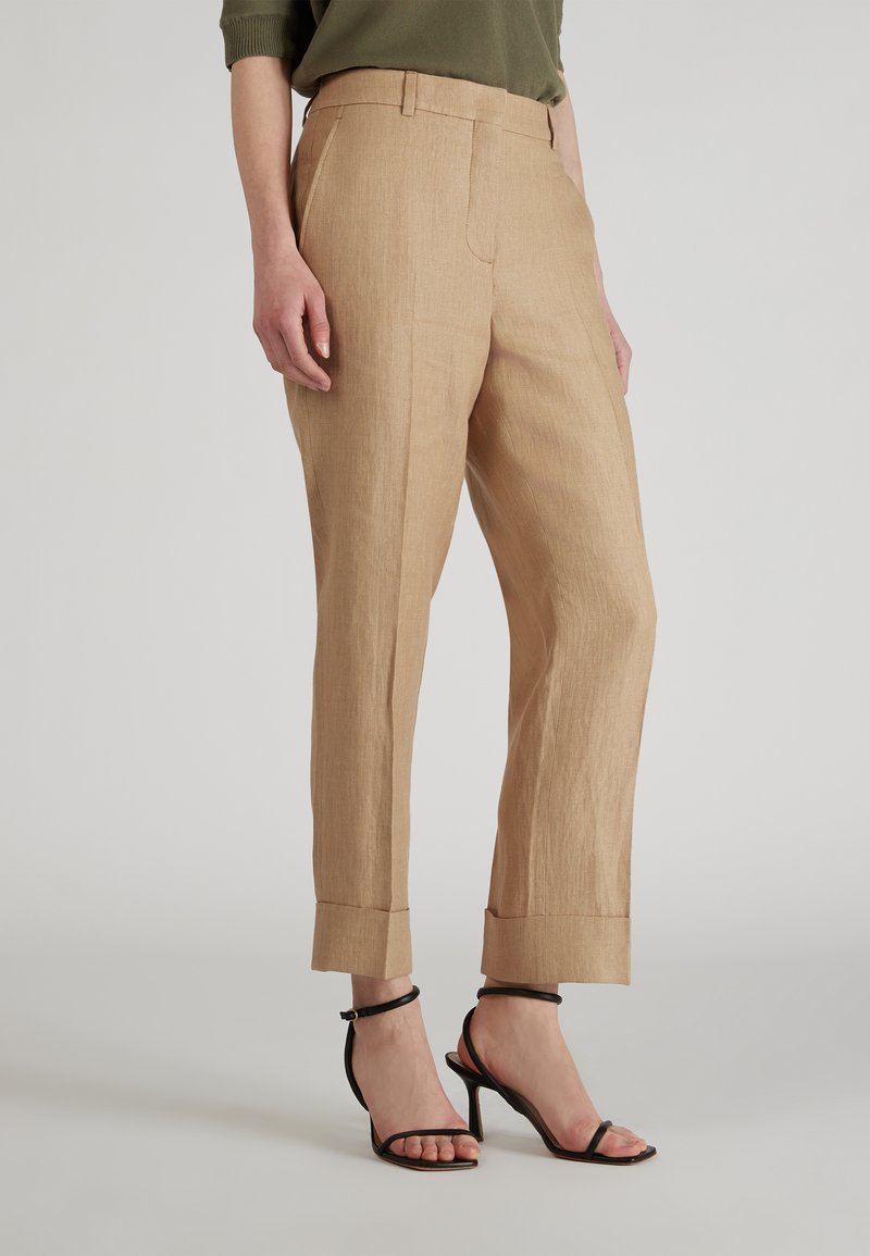 Beżowe lniane spodnie o prostym kroju, z gładką teksturą, przednimi kieszeniami i podwiniętymi nogawkami. Noszone z czarnymi sandałami na obcasie.