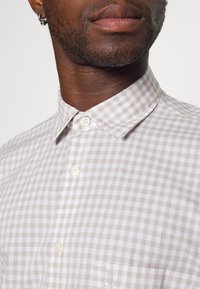 Gros plan d'un homme portant une chemise à carreaux beige et blanche avec une poche et des boucles d'oreilles anneaux argentés.