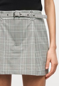 Γυναίκα που φοράει μια ψηλόμεση ανοιχτογκρι καρό φούστα με πιέτες και ιμάντα από το ίδιο ύφασμα, συνδυασμένη με μαύρο τοπ, με το χέρι ακουμπισμένο στο πλάι.