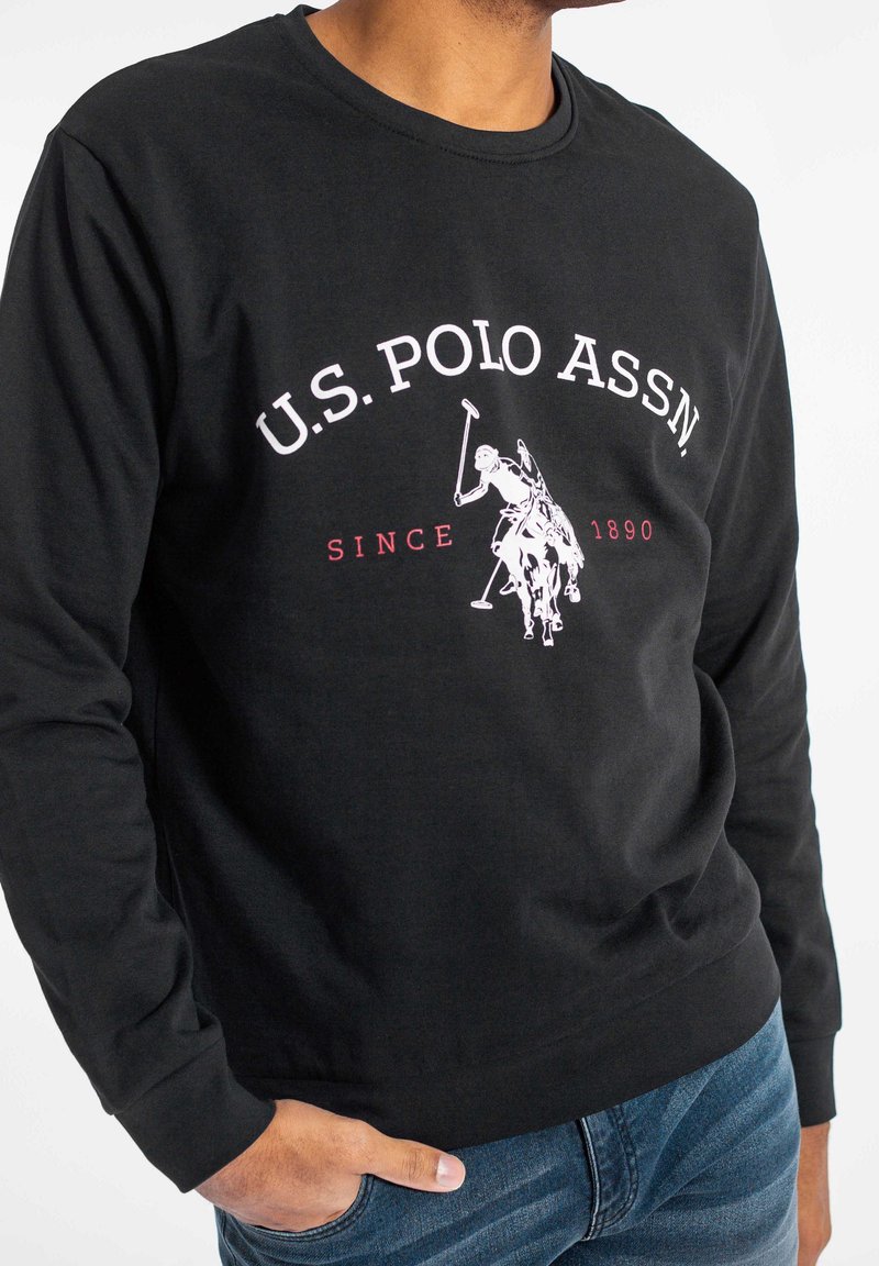 Sudadera negra de manga larga con un logotipo gráfico en el pecho en blanco y rojo, cuello redondo y puños acanalados, combinada con jeans azules.