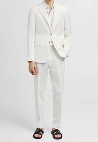 Completo in lino bianco con giacca con un solo petto e pantaloni abbinati, abbinato a sandali neri. Presenta bottoni dorati e una texture liscia.