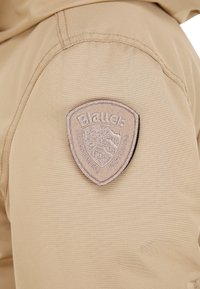 Beige stof med en glat tekstur, prydet med en beige skjoldformet lap med "Blauer" og et bjergdesign syet på venstre side.