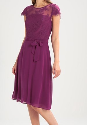 Kobieta w fioletowej sukience do kolan z koronkowym górnym elementem, przejrzystymi krótkimi rękawami i przewiązanym paskiem w talii, stojąca na jednolitym tle.