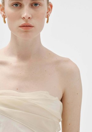 Top strapless de crema transparente con tela suave y fluida drapeada sobre el busto. Pendientes de aro dorados complementan el look. Diseño simple y minimalista.