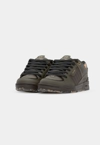 Dark green sneakers with leather and textile upper, black laces, and camouflage patterned sole. Features logo accents on tongue and side.
