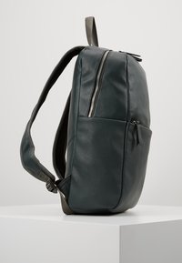 Sac à dos en cuir texturé vert foncé avec des bretelles rembourrées réglables, une poche zippée à l'avant et une fermeture éclair verticale sur le côté du compartiment principal.