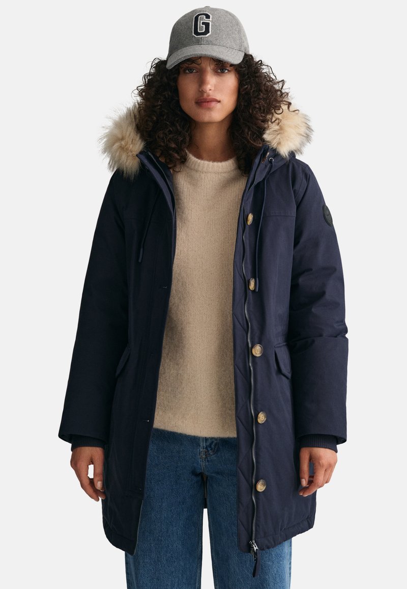 Frau mit einer marineblauen Parka mit Pelzkapuze, beigem Pullover, blauen Jeans und grauem Baseball-Cap mit dem Buchstaben G, steht vor einem schlichten Hintergrund.