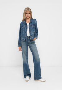 Jeans jakna z svetlo modrim pranjem, zapiranje na gumbe, dva žepna na prsih in sproščen kroj, v kombinaciji z usklajenimi širokimi hlačami in belimi supergami.