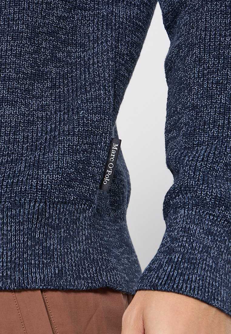Marineblauer Strickpullover mit strukturierter Oberfläche, gerippten Bündchen und einem sichtbaren Etikett mit der Aufschrift "Marc O'Polo". Nahaufnahme des Ärmels.