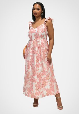 Frau trägt ein ärmelloses, pink-goldenes Maxikleid mit Blumenmuster und Rüschen-Trägern, steht mit einer Hand in der Tasche und trägt goldene High Heels.