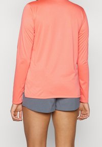 Koraalkleurig shirt met lange mouwen van een textuurstof, met zijsplitten en een ronde onderrand. Gecombineerd met grijze shorts.