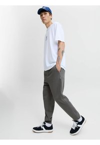 Hvid t-shirt med korte ærmer, grå bukser med sidelommer og sorte sneakers med hvide detaljer. Blå kasket på hovedet, stående stilling.
