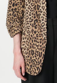 Leopardmönstrad kimono-jacka med vida ärmar, beige och bruna färger, med en mjuk textur och avslappnad passform. Svarta byxor syns.